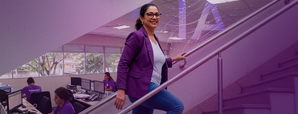 Mulher de cabelo preso, óculos e blazer roxo está subindo escadas enquanto sorri para a câmera
